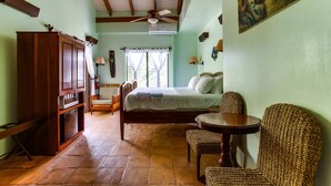 A bedroom with a bed, a wooden wardrobe, a wicker chair, a small round table, and a ceiling fan. - TBS 33 Beachside-Resort Style with Pool, 2 Bedroom (Placencia)