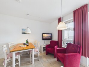 A modern living room with a wooden dining table, white chairs, a red sofa, and a television on the wall. - Nordland Apartments - Langeneß Apartment - Right on the water—you can't get any closer than this! (Wyk auf Föhr)