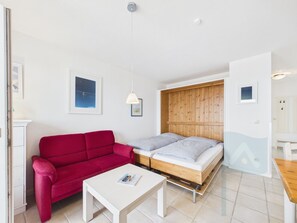 A compact room with a red sofa, a single bed, a white table, and a wooden headboard.