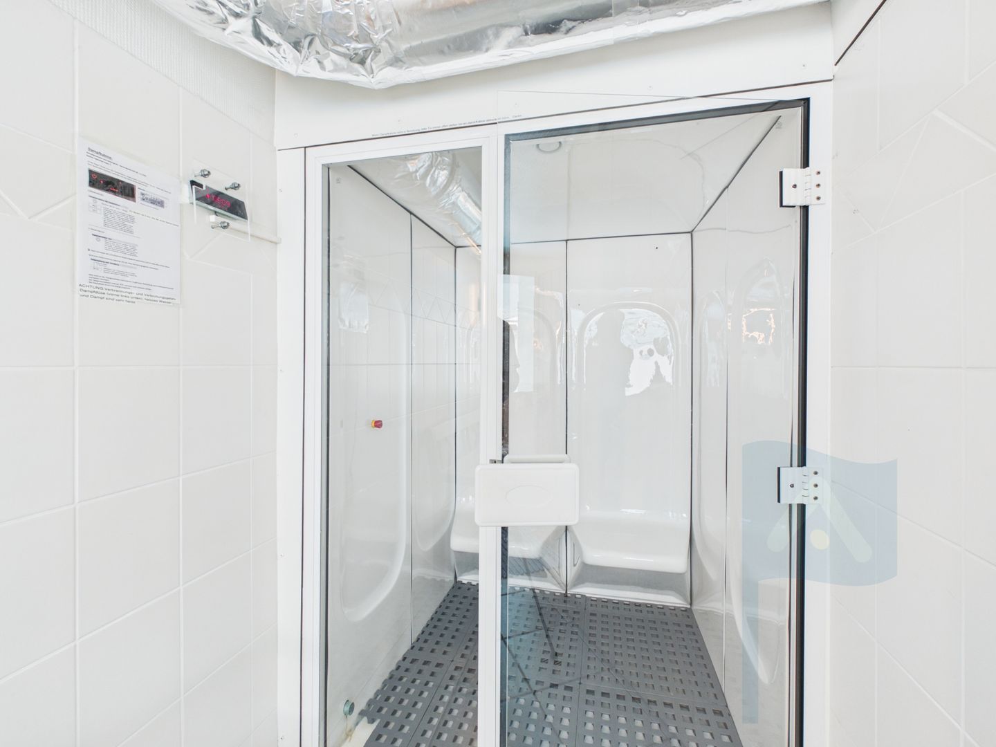 A glass-enclosed shower stall with a metal grate floor, white tiled walls, and a visible electrical outlet on the wall.