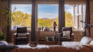 Un salon chaleureux avec deux fauteuils en cuir, un canapé, une table avec des livres et des objets décoratifs, un télescope, et une vue sur les arbres et les montagnes aux couleurs automnales, visible à travers de grandes fenêtres.