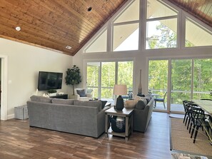 A spacious living room with a large gray sectional sofa, a flat-screen TV, and a large window offering a view of trees.