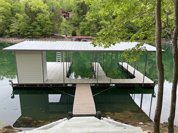 A dock extending into a calm lake, surrounded by trees and a house in the background.