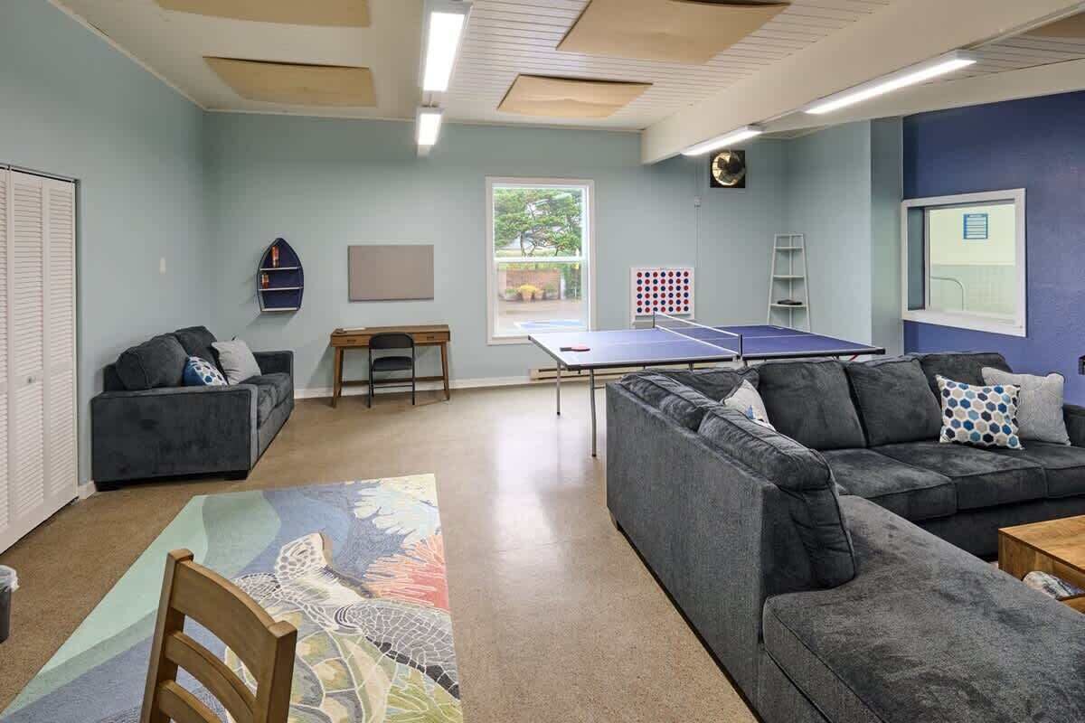 Una habitación con dos sofás grises, una mesa de ping pong, una mesita, una silla, una ventana y una puerta.