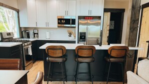 Una cucina moderna con mobili bianchi, elettrodomestici in acciaio inossidabile e pavimento in legno. Sono presenti tre sgabelli da bar e un tavolo da pranzo con sedie.