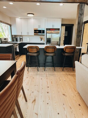 A modern kitchen with white cabinets, stainless steel appliances, and a wooden floor. There are three bar stools and a dining table with chairs. - Lake Chatuge Waterfront Retreat_Dock+Hot Tub+Fire Pit_Sleeps 12 (Hiawassee)
