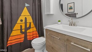 A bathroom with a shower curtain featuring a geometric design, a toilet, a sink with a mirror, and a small potted plant. - Chaparral Charmer | 3BR | Pool | Bikes | King bed! (Scottsdale)