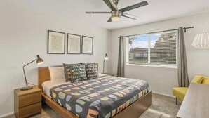 A bedroom with a bed, a nightstand, a desk, a chair, a ceiling fan, and a window with blinds. - Chaparral Charmer | 3BR | Pool | Bikes | King bed! (Scottsdale)