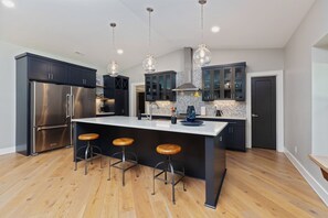 Een moderne keuken met donkere kasten, een roestvrijstalen koelkast, een groot eiland met krukken, hanglampen en een backsplash met een herringbone-patroon.