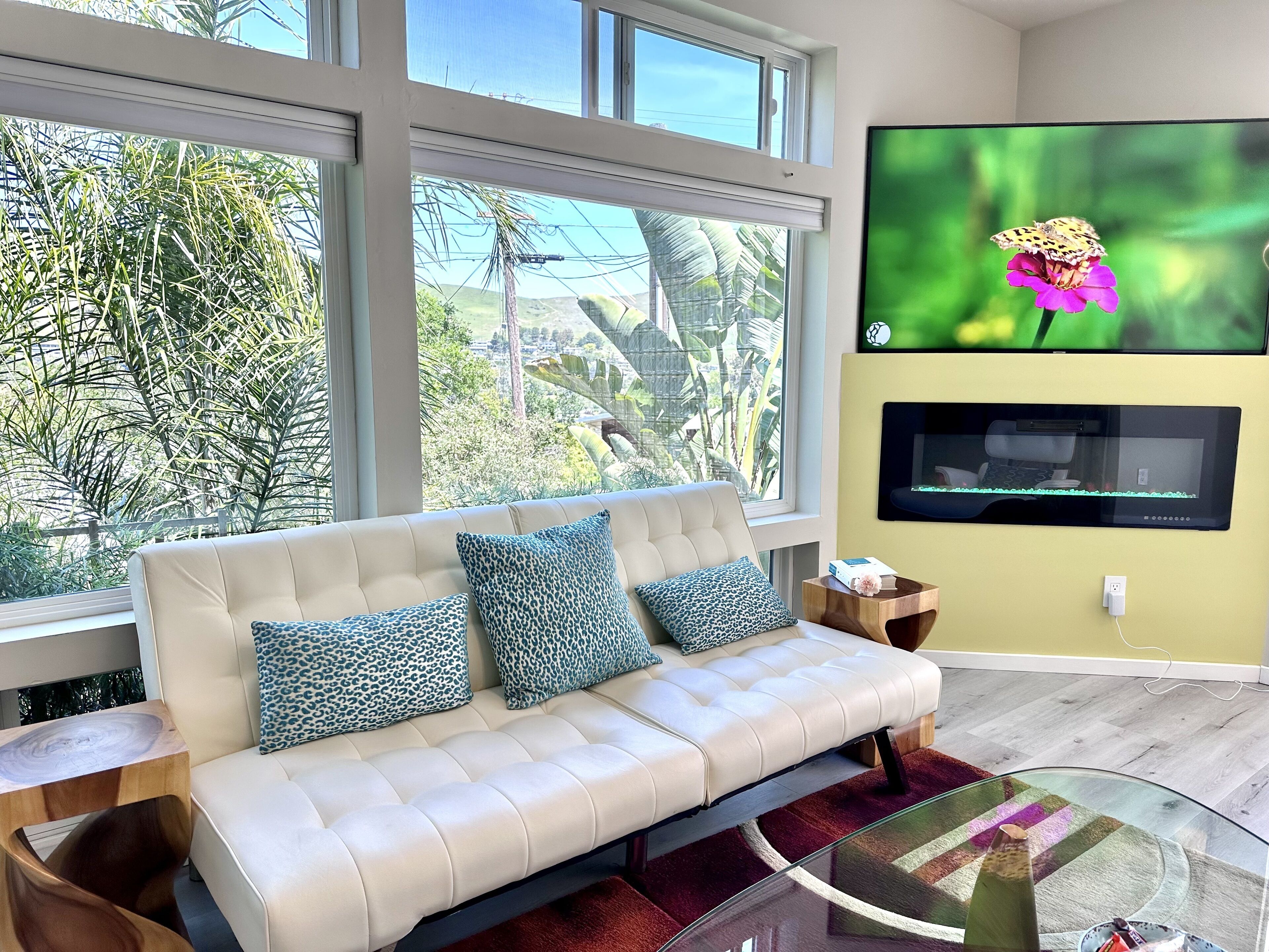 Een moderne woonkamer met een beige bank, kussens met luipaardprint, een glazen salontafel en een flatscreen televisie aan de muur bevestigd.