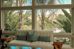 A room with a sofa facing large windows, a wooden side table, and a view of tropical vegetation.