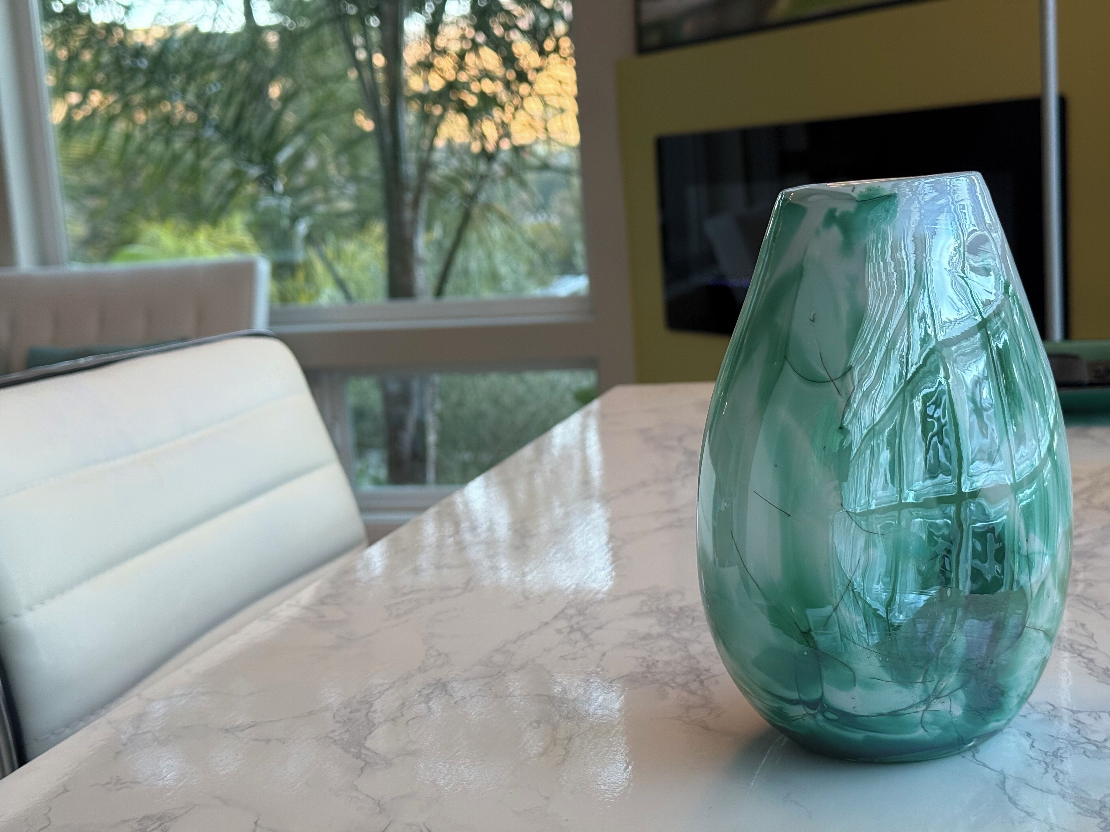 A green glass vase on a marble table in a room with a white chair and a window with a view of trees.