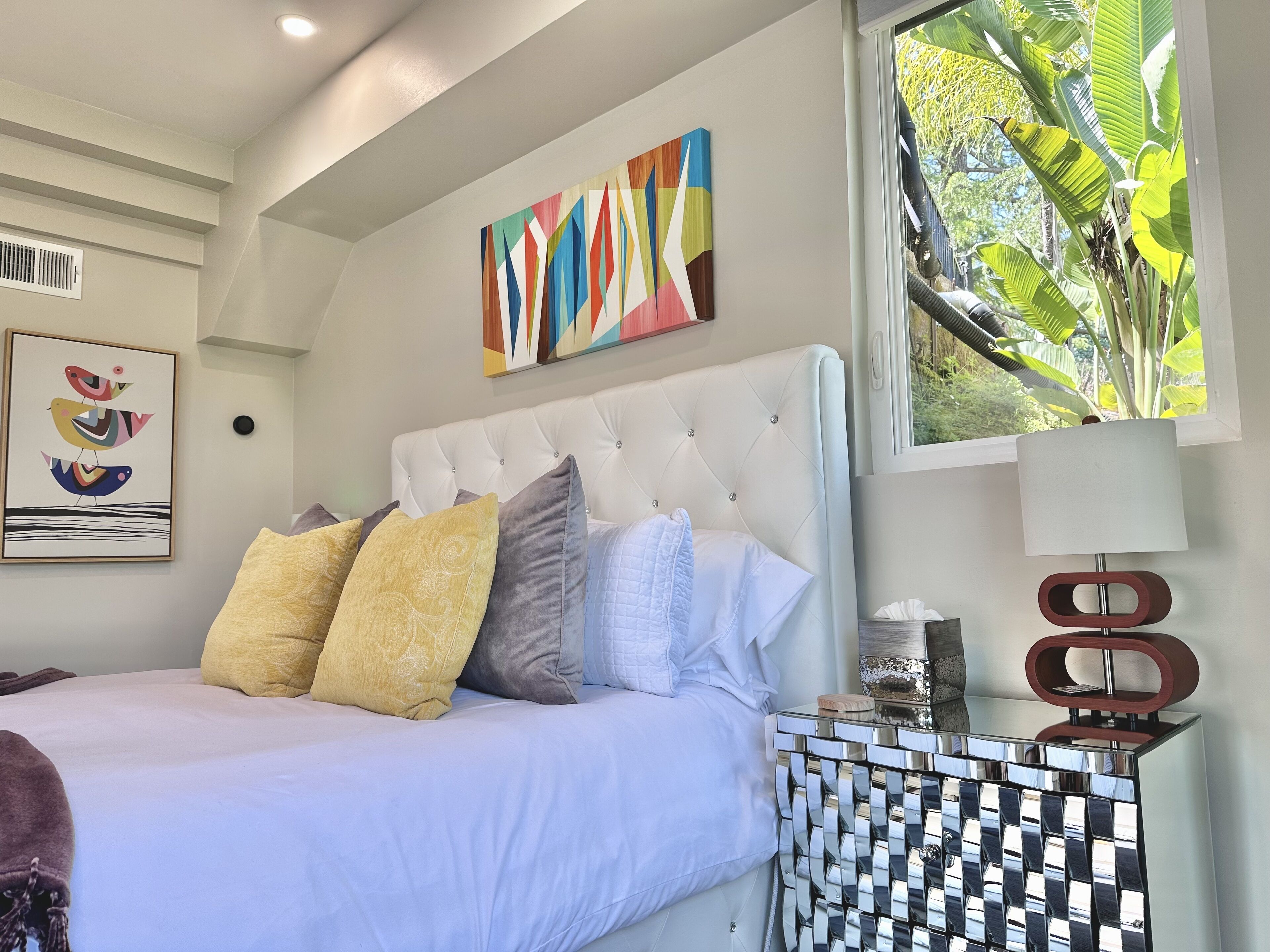 A bedroom with a white bed, multiple pillows, a colorful abstract painting, a framed artwork of birds, a window with a view of greenery, and a modern side table with a lamp.
