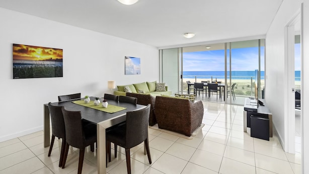 A modern living room with a dining area, large windows offering a view of the beach, and a balcony with a table and chairs. - Kirra Beach 506 Pure Ocean Views (Coolangatta)