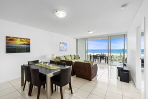 A modern living room with a dining area, large windows offering a view of the beach, and a balcony with a table and chairs.