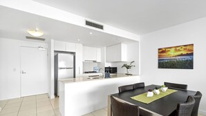 A modern kitchen with white cabinets, a central island, and a dining area with wicker chairs. - Kirra Beach 506 Pure Ocean Views (Coolangatta)
