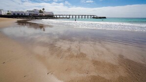 A beach with a pier extending into the sea, a clear sky, and buildings in the background. - Lanzahost Villa Mimosa (Punta Mujeres)