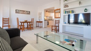 Un soggiorno moderno con un tavolino in vetro, sedie in legno, una divano, un televisore e una cucina sullo sfondo.