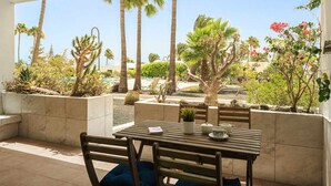 Una terrazza esterna con tavolo e sedie in legno, circondata da fioriere con diverse piante e alberi, e vista su un'area piscina.