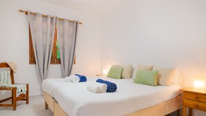 Un letto rifatto con cura, asciugamani arrotolati, un comodino in legno, una sedia con cuscino a righe e una finestra con tende.
