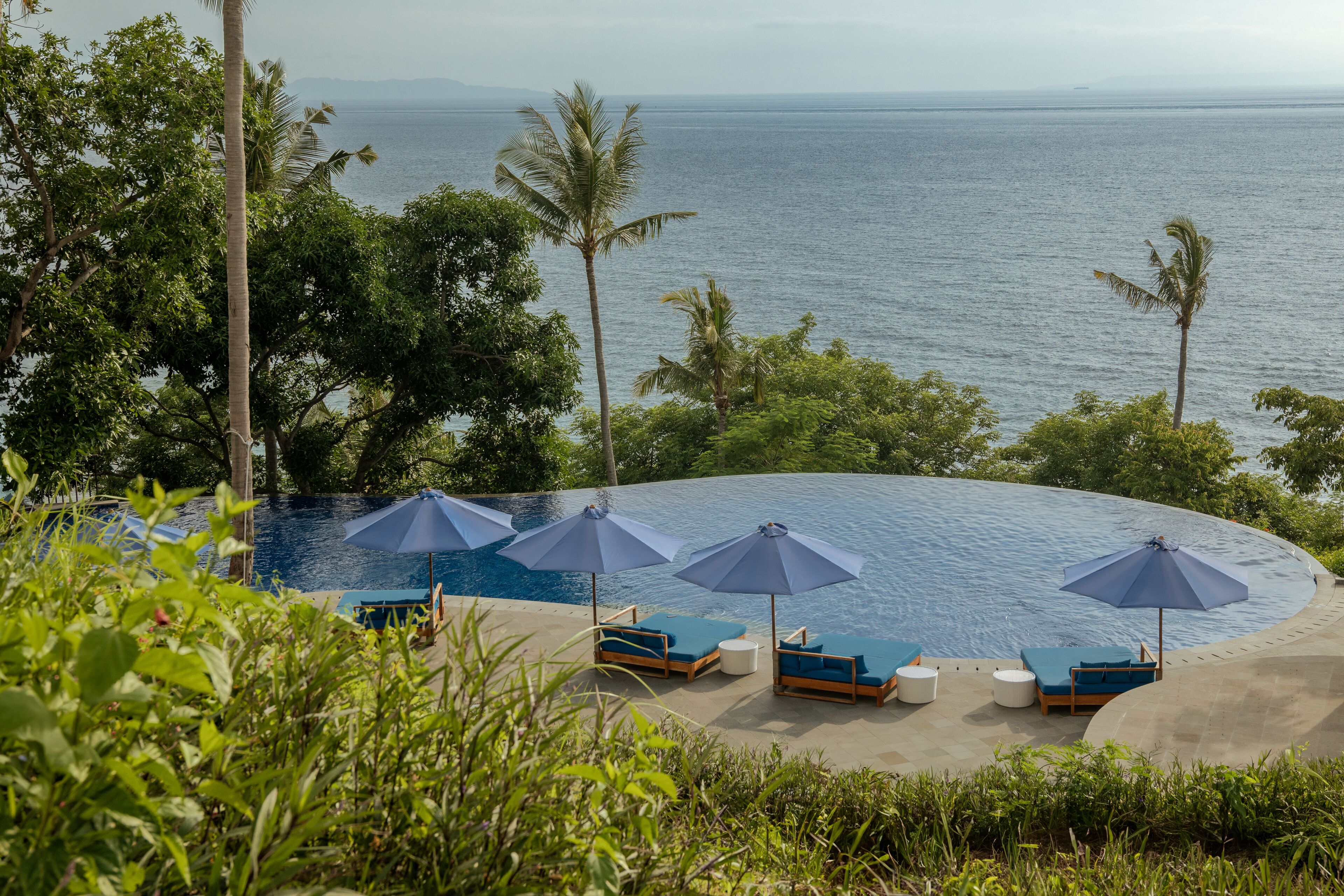A poolside area with lounge chairs and umbrellas, surrounded by palm trees and overlooking the sea.