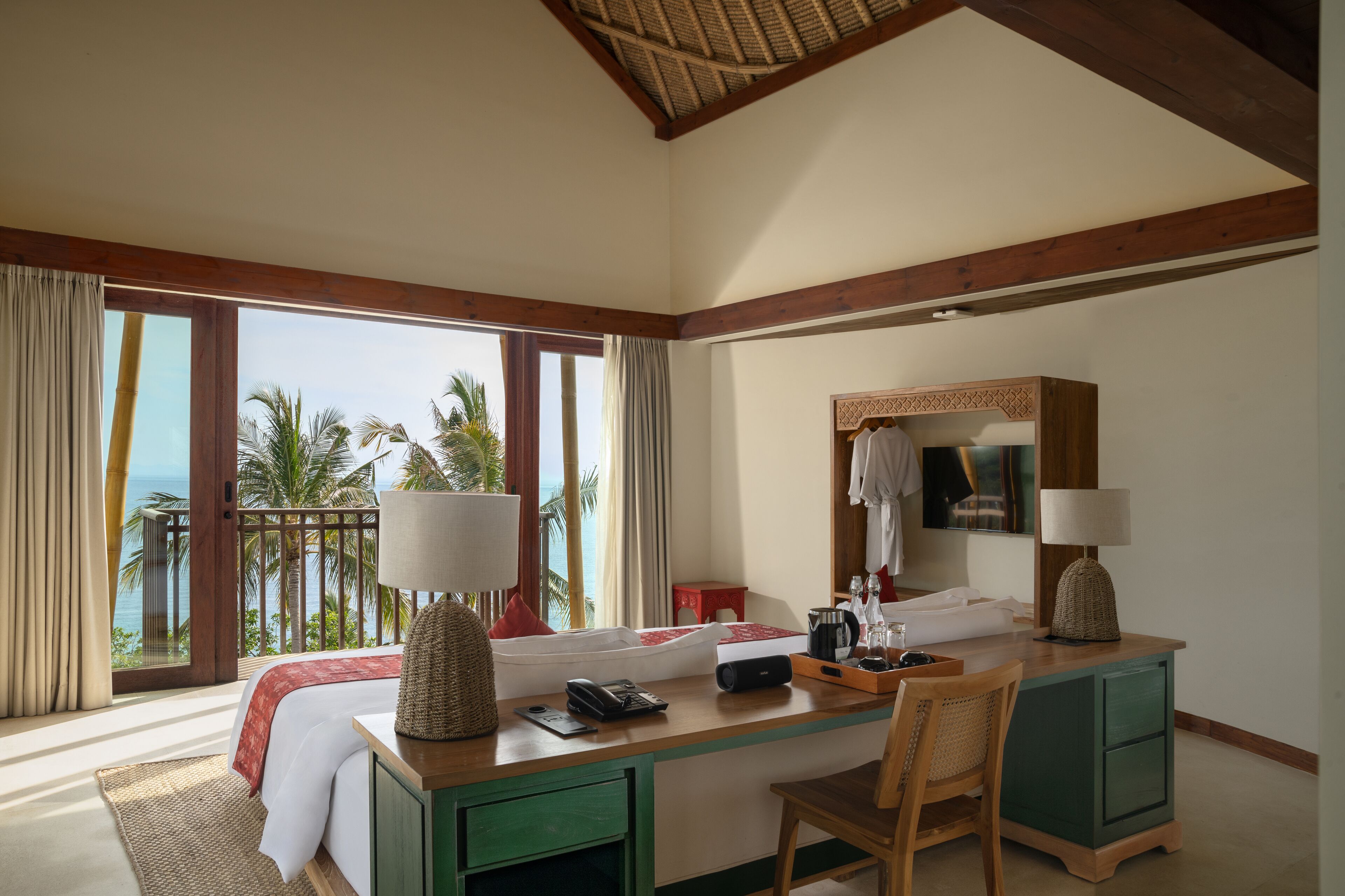 A hotel room with a large bed, a desk with a chair, a mirror, and a view of palm trees and the ocean.