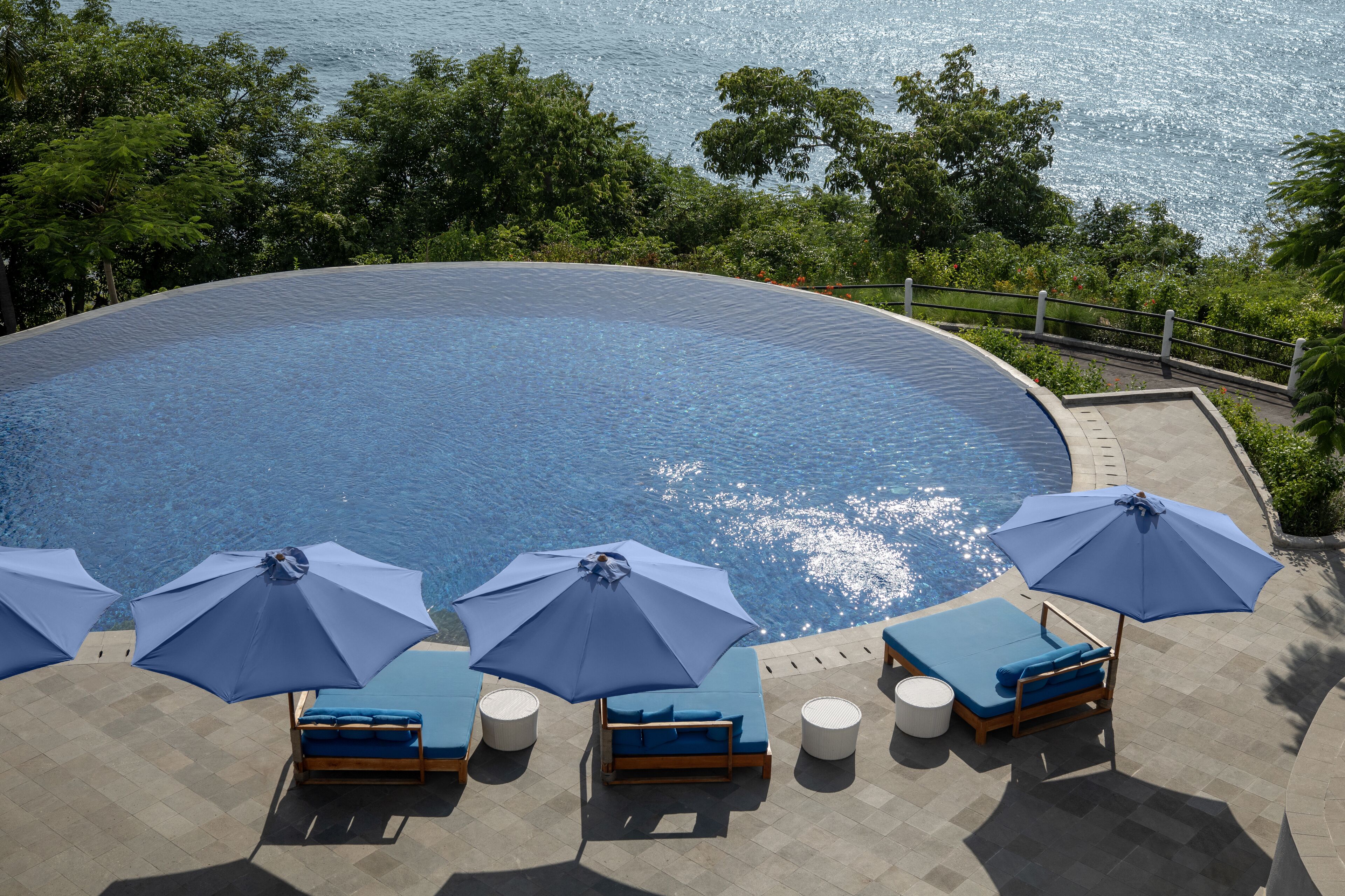 Un área junto a la piscina con sillones de descanso y sombrillas, rodeada de vegetación y con vista a un cuerpo de agua.