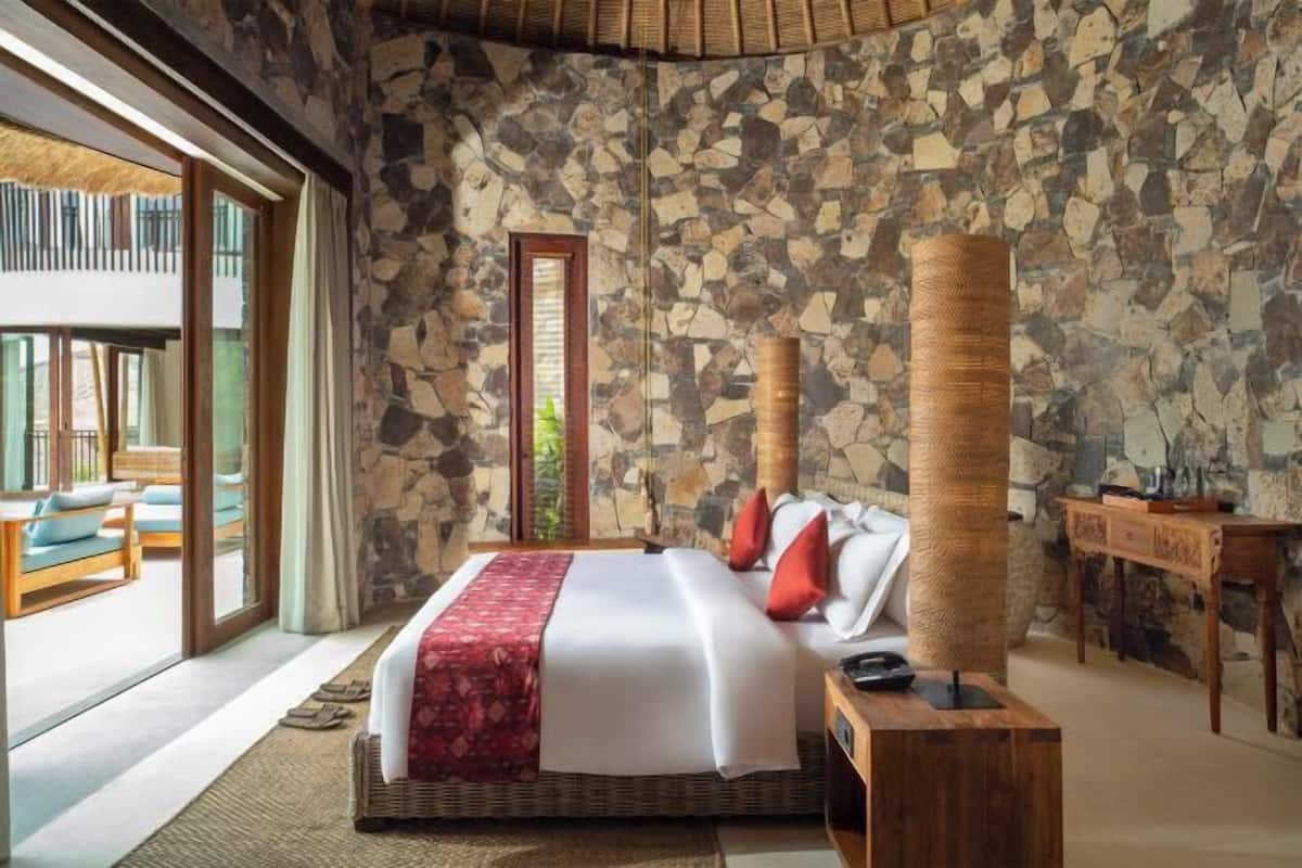 A bedroom with a stone wall, a bed with white bedding and red pillows, a wooden nightstand, and a large window.