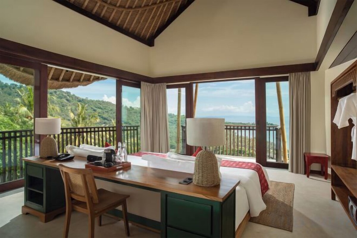 A room with a desk, chair, bed, and large windows overlooking a scenic view.