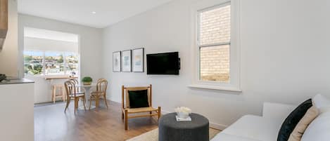 A modern living room with a white sofa, a round ottoman, a dining area with a table and chairs, and a TV mounted on the wall.