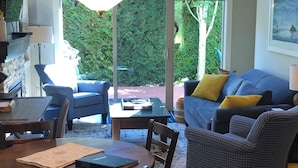 Et spiseområde med et bord og stole, en sofa med puder, og et stort vindue med udsigt til grøn natur.