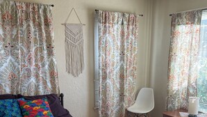 Une chambre avec un lit violet, des coussins colorés, des rideaux à motifs, une chaise blanche et une petite table avec une lampe.