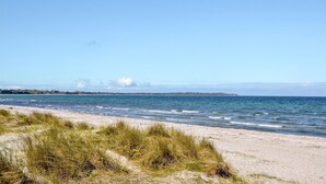 A beach with sand and grass in the foreground, a calm sea, and a distant shoreline with buildings.