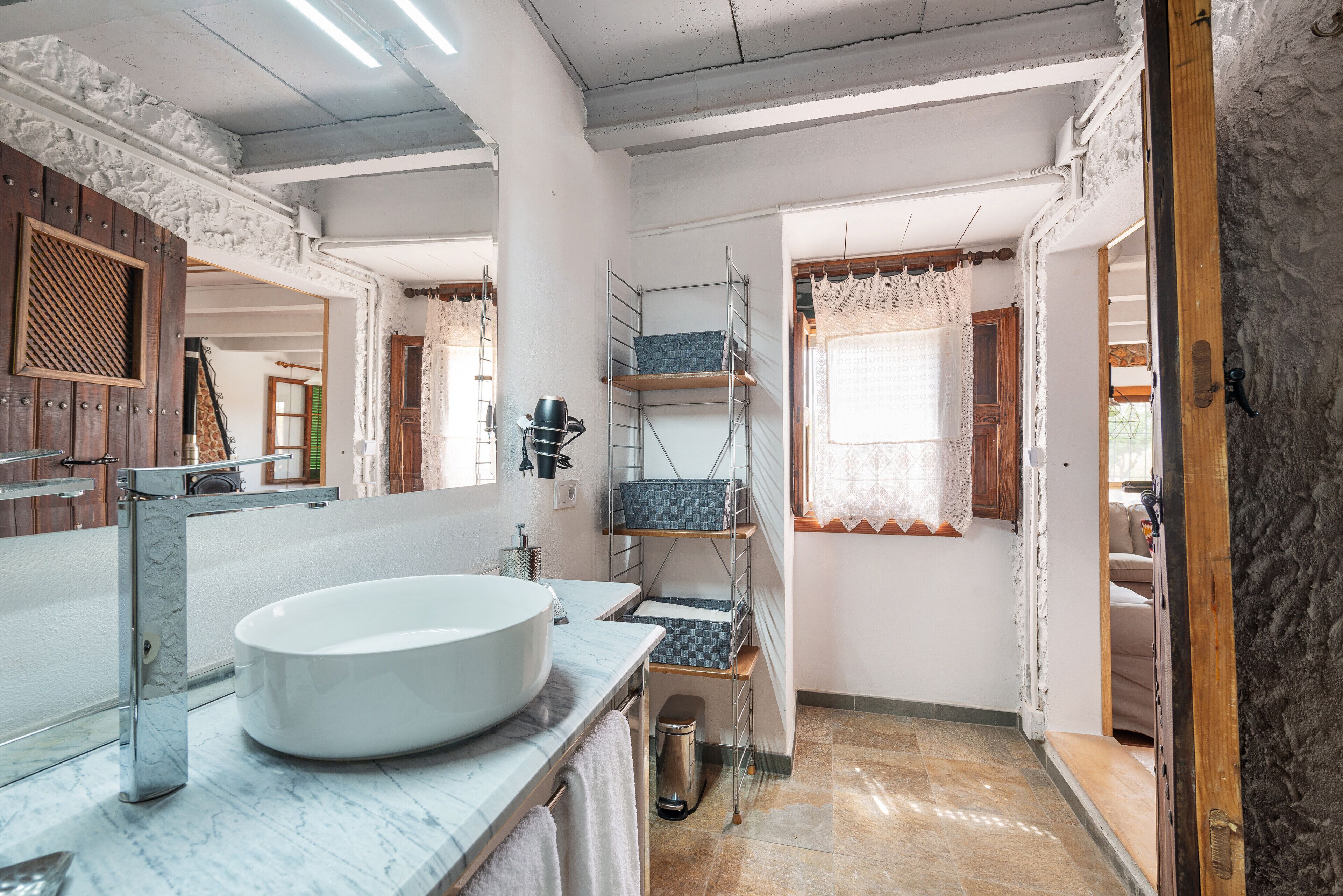 A bathroom with a large white sink, a mirror, and a shower area. There are shelves with storage boxes and a small trash bin.