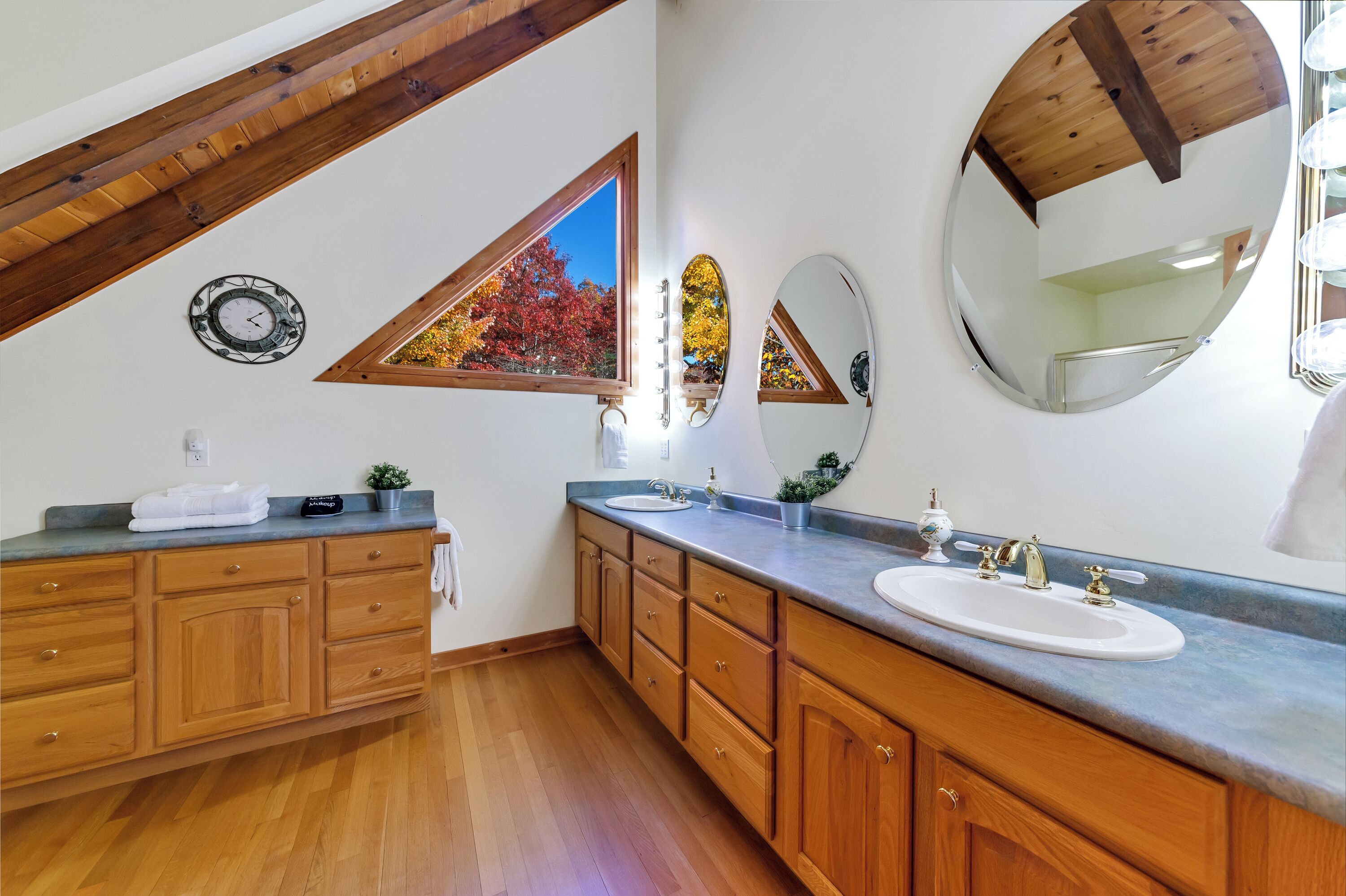 A modern bathroom with wooden cabinets, a large mirror, and a window with autumn leaves.
