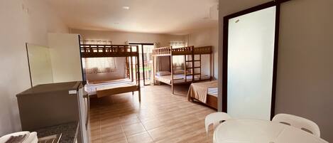 Quarto de hotel com uma cama de madeira beliche, uma escrivaninha com cadeira, uma mesa de jantar com cadeiras e uma janela grande.