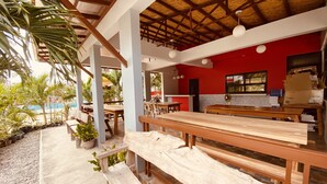An outdoor dining area with wooden benches and tables, surrounded by potted plants and a pool.