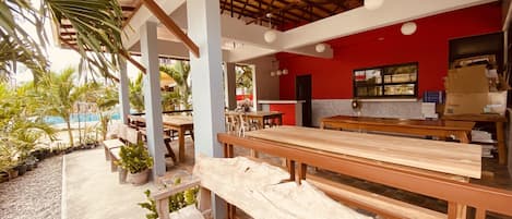 An outdoor dining area with wooden benches and tables, surrounded by potted plants and a pool.