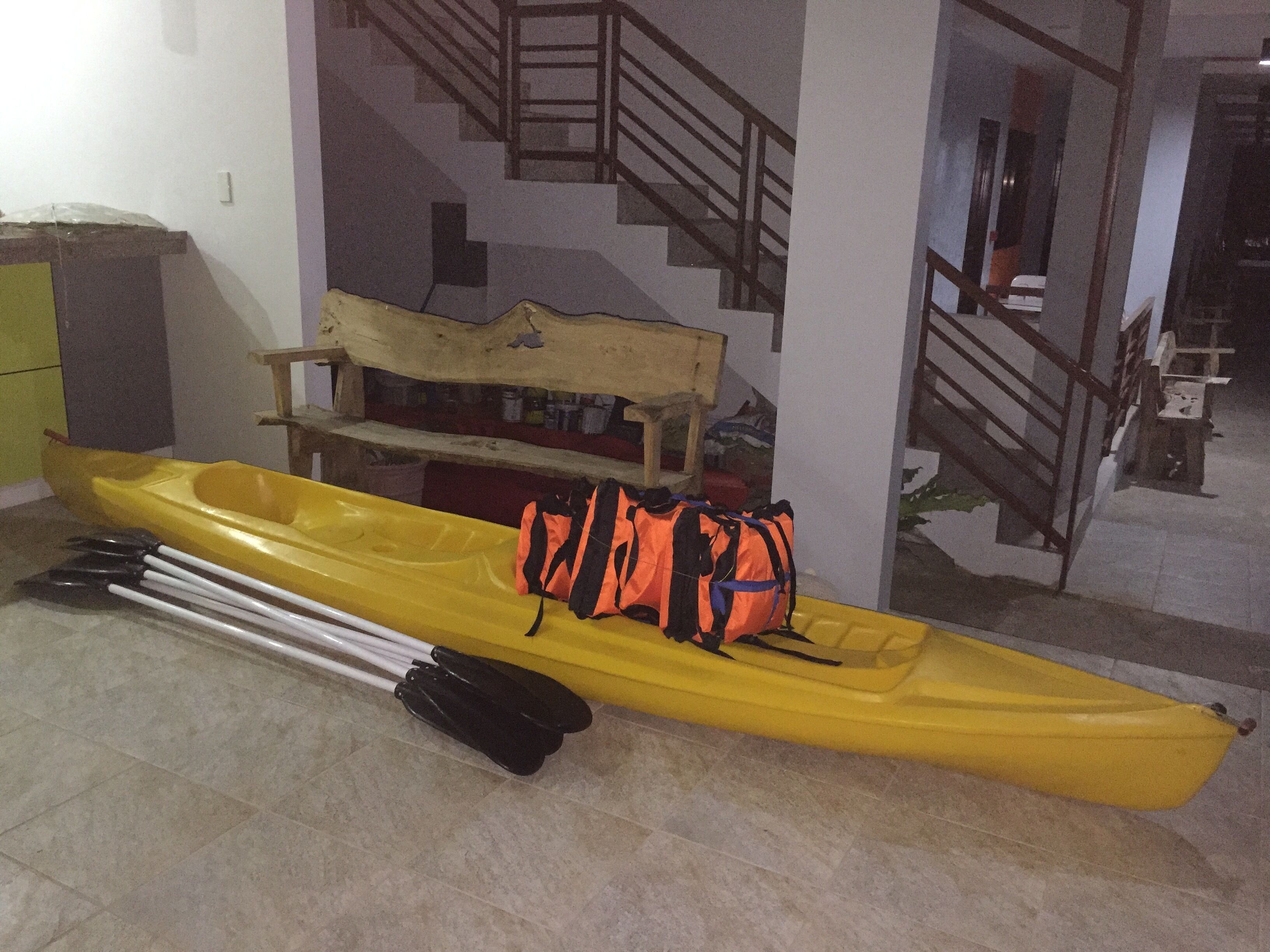 Un kayak amarillo con remos y un chaleco salvavidas naranja sobre un suelo de baldosas.