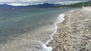 A pebble beach with clear water, a boat, and mountains in the background.