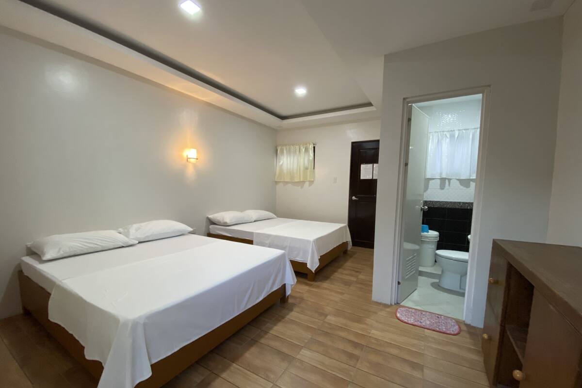 A hotel room with two beds, a wooden floor, a door leading to a bathroom, and a wall-mounted light fixture.