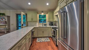 A kitchen with a stainless steel refrigerator, wooden cabinets, a marble countertop, and a view of trees through a window. - Cozy Clayton Cabin w/ Deck & Mountain Views! (Clayton)