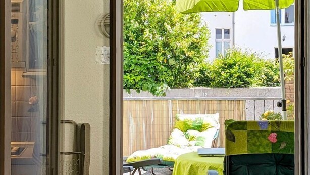 Eine Glastür öffnet sich zu einer Außenveranda mit einem grünen Sonnenschirm, einem Tisch mit gelber Tischdecke und einem gepolsterten Stuhl.