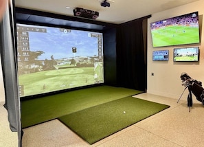 A room with a large screen displaying a golf course, a golf bag, and a projector mounted on the ceiling.