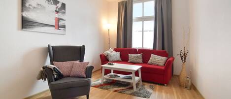 Un salón con un sillón gris, un sofá rojo, una mesa de centro blanca y un cuadro de un faro.