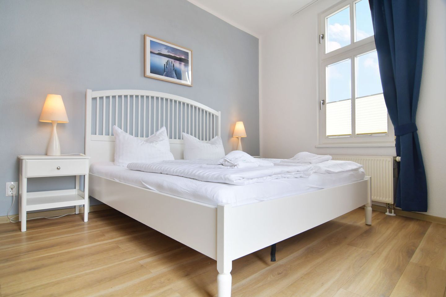 A neatly made bed with white bedding, two bedside tables with lamps, a framed picture on the wall, and a window with curtains.