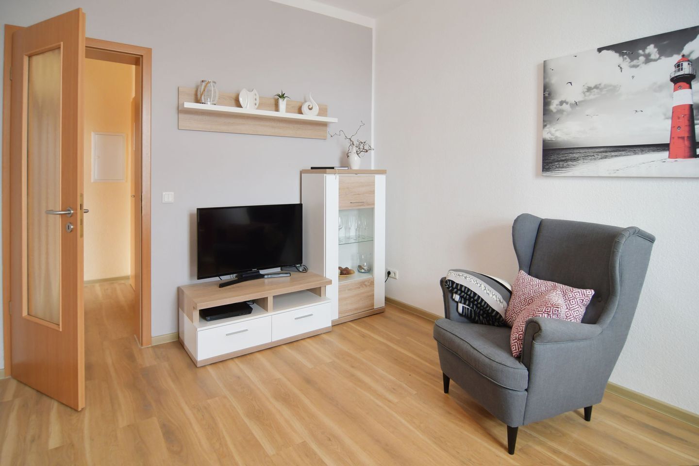 A living room with a TV on a wooden stand, a grey armchair, and a shelf with decorative items.