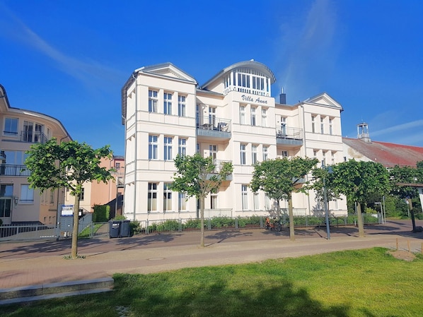 Een meerlaags gebouw met balkons, genaamd "Villa Anna," omringd door bomen en een heldere lucht.