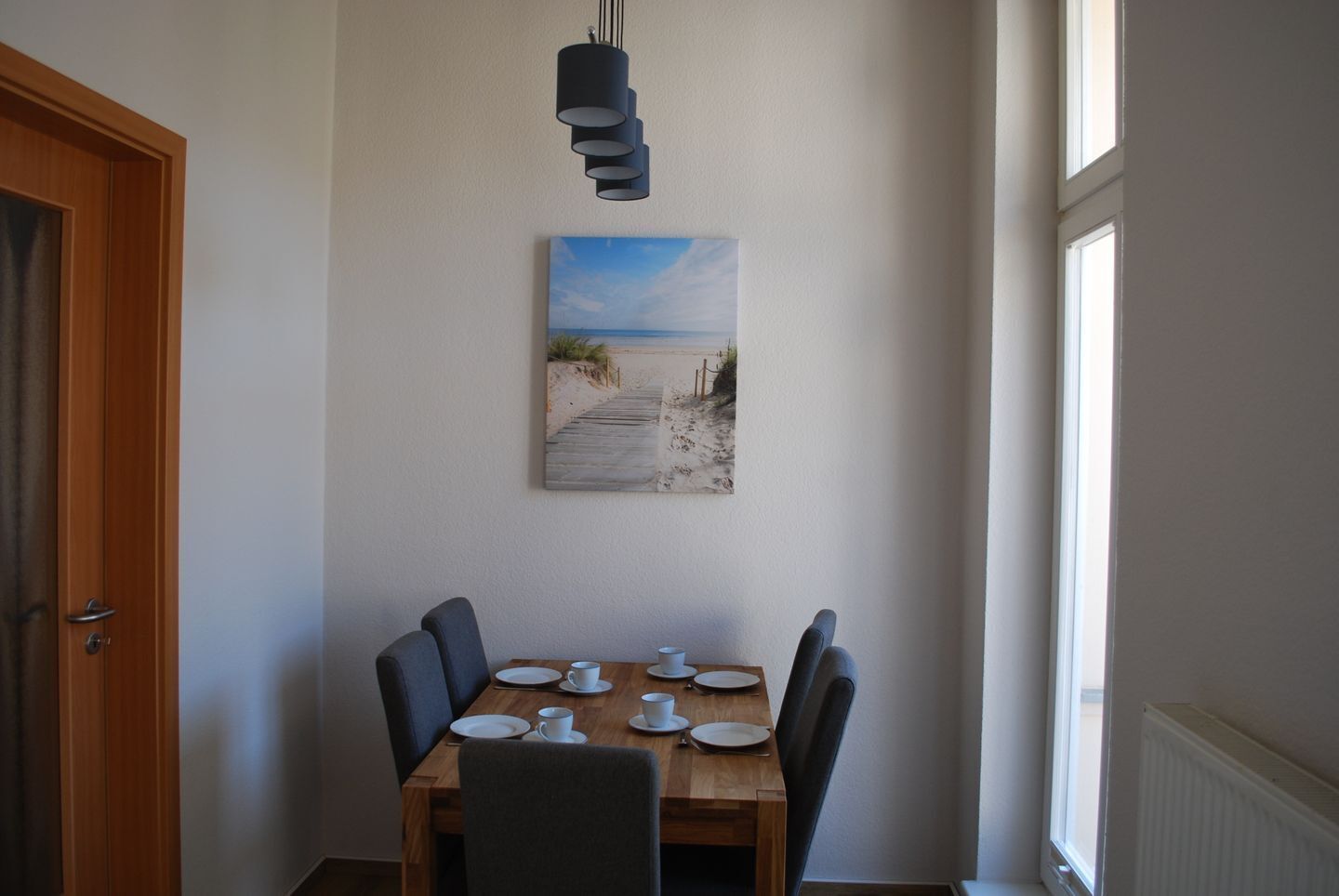A dining area with a wooden table set for four, a painting of a beach scene, and a door to the left.