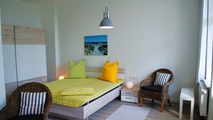 A modern bedroom with a bed, two wicker chairs, a nightstand, a lamp, and a painting on the wall. - Haus Wanda Appartement 4a - Haus Wanda (Heringsdorf)